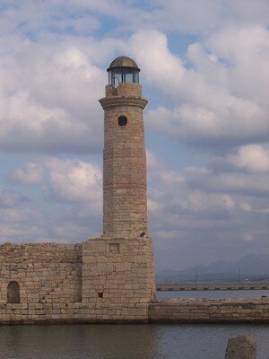 The historic Venetian Lighthouse in Rethymno, Crete, standing on a stone pier against a backdrop of cloudy skies and calm waters.