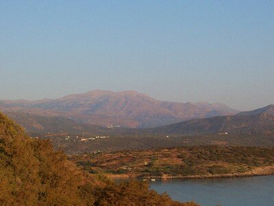 A scenic view of rolling hills and rugged mountains in Crete, bathed in warm golden light. The foreground features lush vegetation and a calm coastal inlet, creating a tranquil and picturesque Mediterranean landscape.